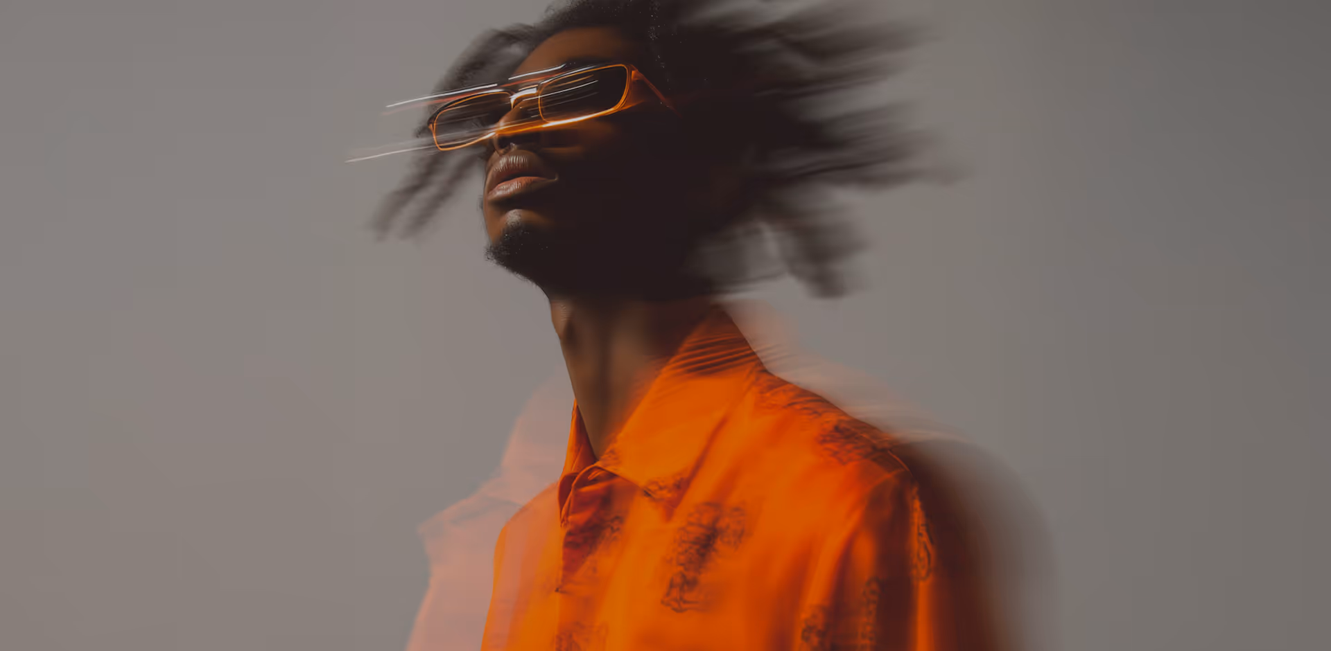 Portrait of a man in an orange patterned shirt and orange-tinted sunglasses with blurred motion effect on hair and shirt against a gray background.